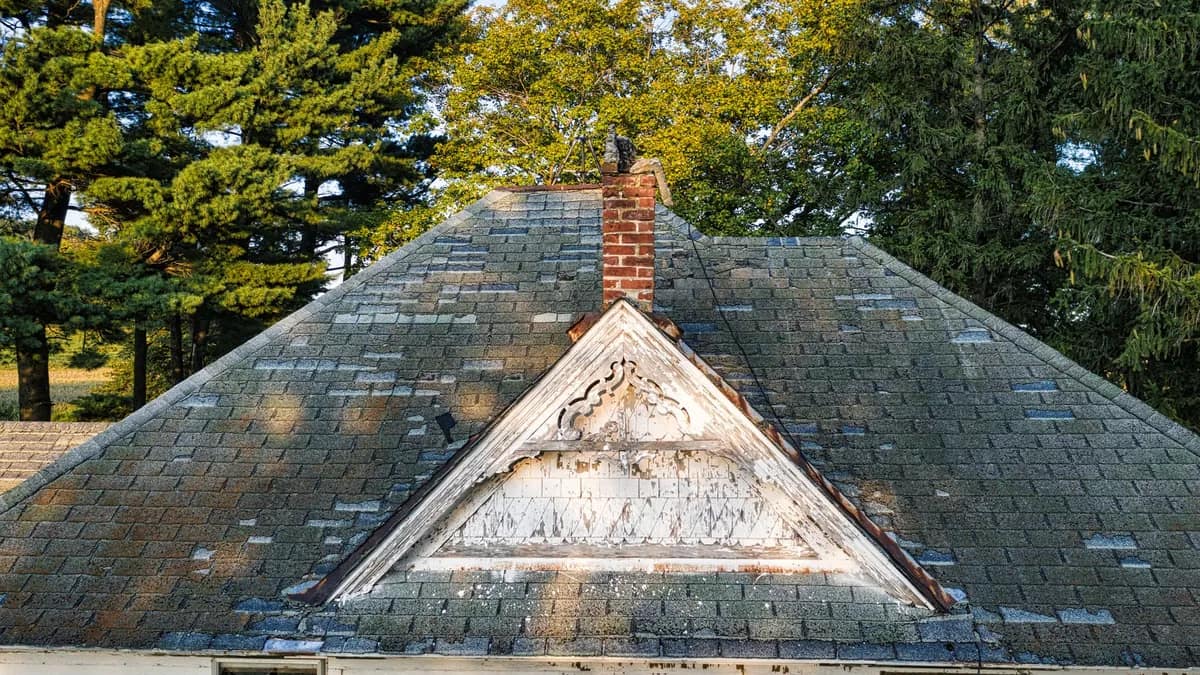 Heavily weathered shingle roof and red brick chimney on an old white clapboard farmhouse