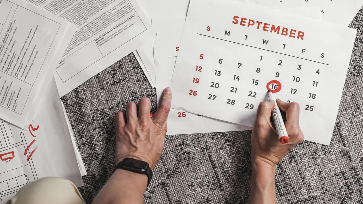A hand circling a date on a wall calendar laid over a stack of paperwork