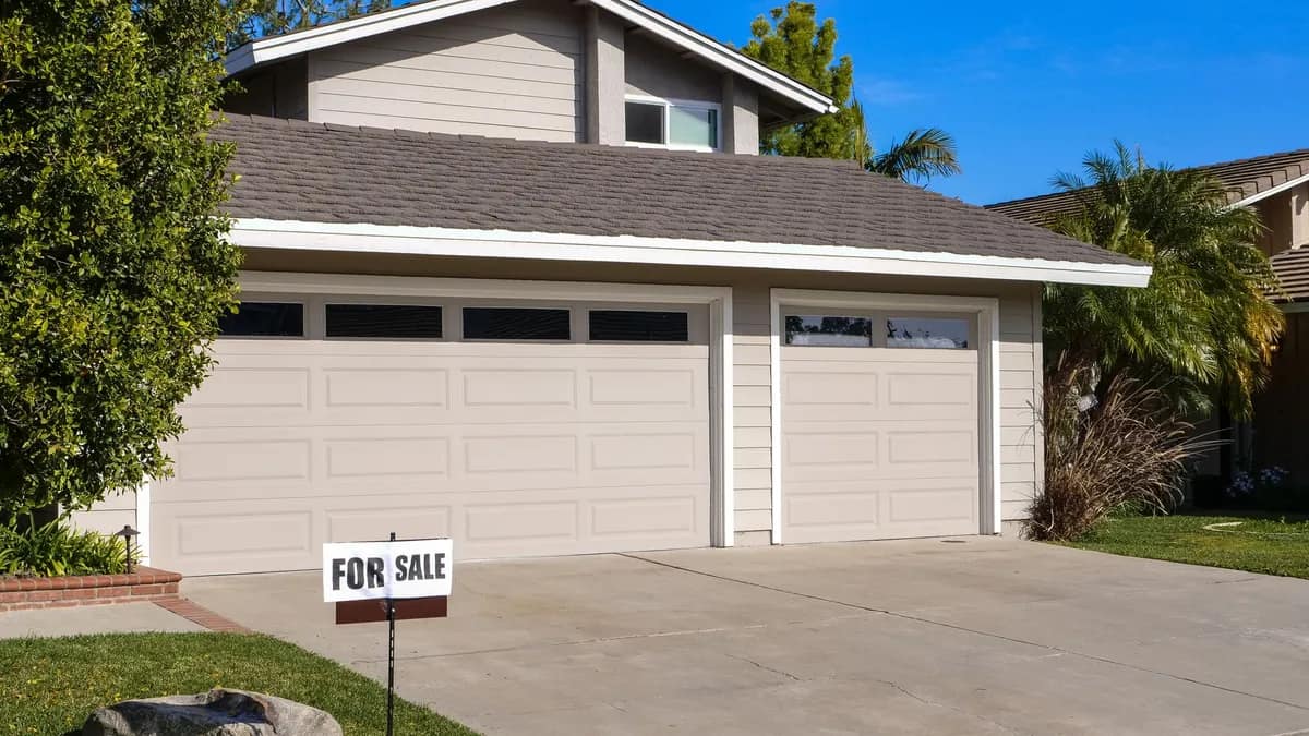 A For Sale sign in the front yard of a suburban home with a shingle roof and two-car garage