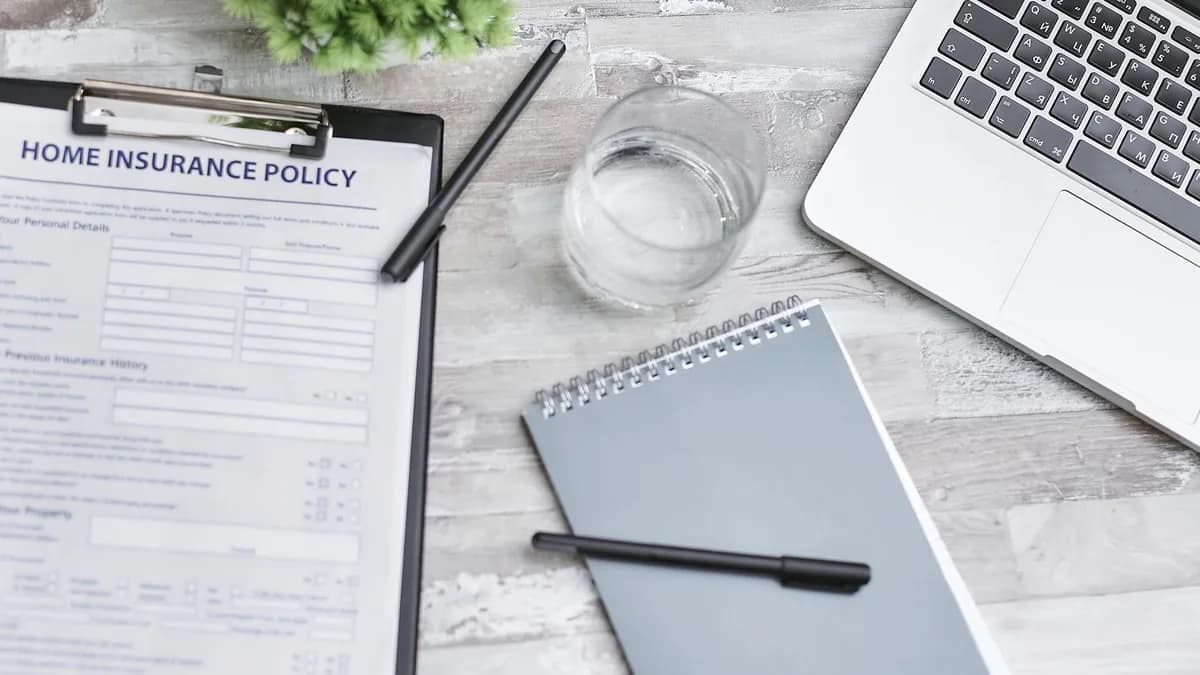 A home insurance policy form on a clipboard beside a laptop, notepad and pen