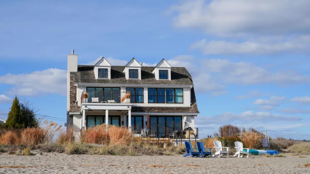 An older weathered beach house with dormers and a dark shingle roof standing on open sand