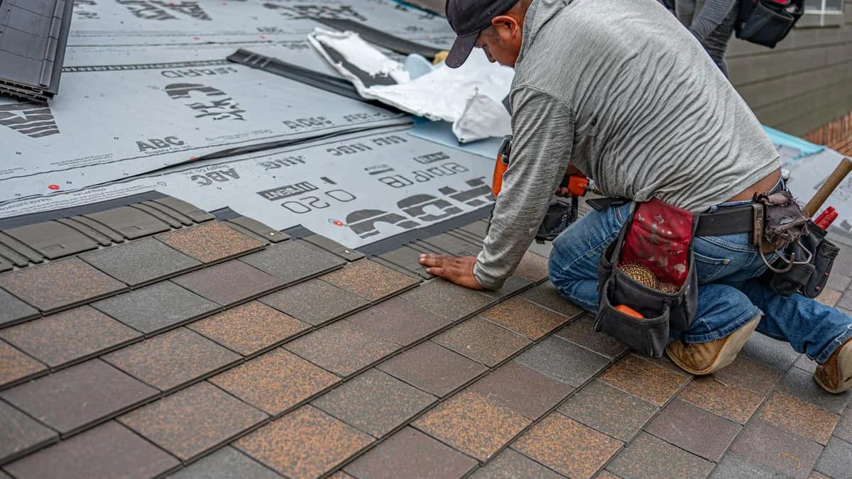 A roofer smoothing synthetic underlayment across a bare roof deck before shingles go down